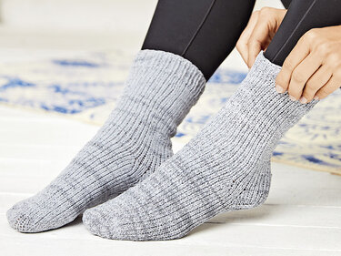 Füße in grauen, gerippten Stricksocken mit schwarzen Leggings; eine Hand hält den Schaft der Socke.