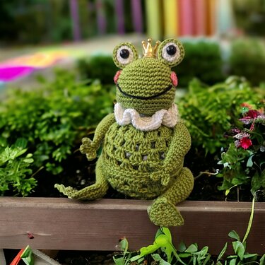 Gehäkelter grüner Frosch mit goldener Krone und weißem Rüschenkragen, sitzend auf einer Holzkante vor Moos und kleinen Blüten