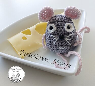 Gehäkelte graue Amigurumi-Maus mit rosa Ohren und Beinen neben einer Käsescheibe auf einer weißen Schale.