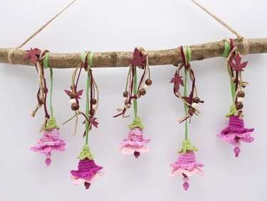 Fünf gehäkelte Blumenanhänger in Rosa und Lila an einem Ast mit Perlen und Bändern