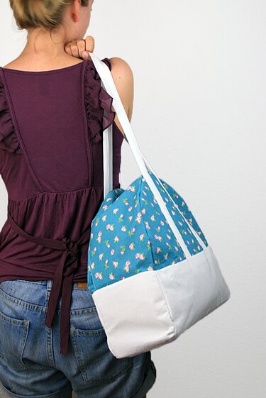 Rückenansicht einer Person in burgunderfarbenem Top mit einem Shopper aus blauem Blumenstoff und hellem, eckigem Boden über der Schulter