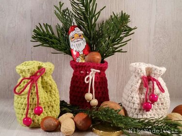 Drei gehäkelte Geschenkbeutel in Gelb, Rot und Weiß mit Kordel und Perlen, arrangiert mit Tannengrün, Nüssen und einer Schokoladenfigur