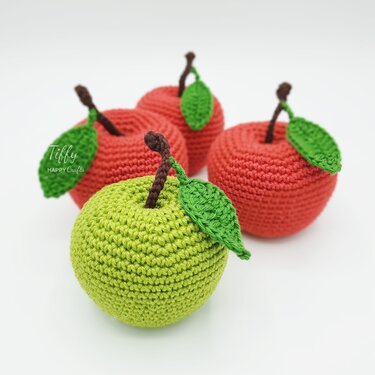 Four crocheted apples, one green in front and three red behind, each with a brown stem and green leaf