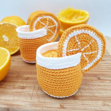 Pair of orange-and-white crocheted cup-like containers with flat orange slice motifs on a wooden cutting board.