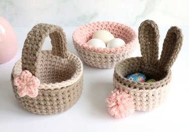 Three crocheted baskets and a bowl in beige, brown, and pink: a handled basket with a flower, an egg bowl, and a bunny basket with eggs.