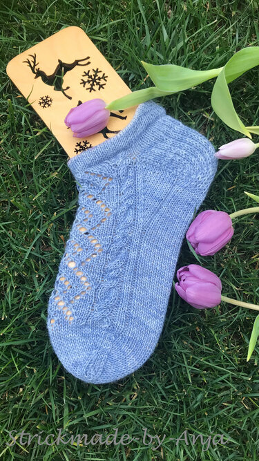 Hellblaue gestrickte Kurzsocke mit Loch- und Zopfmuster auf Gras neben lila Tulpen und Sockenbrett