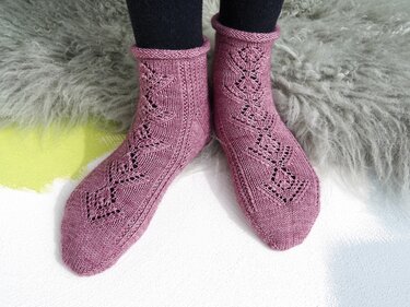 Pair of mauve knitted ankle socks with leaf lace motif and rolled cuffs on a grey faux-fur rug.
