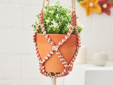 Nahaufnahme zweifarbiger Makramee-Blumenampel in Creme und Rosa mit Tontopf und grüner Pflanze.