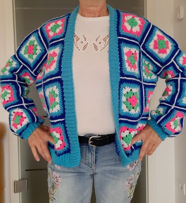 Person trägt offene, bunte Granny‑Square‑Jacke in Blau, Pink, Grün und Weiß über weißem T‑Shirt und Jeans.