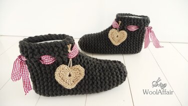 Zwei dunkelgraue, gestrickte Hausschuhe mit rot-weiß karierten Bändern und beigen Häkelherzen auf hellem Boden.