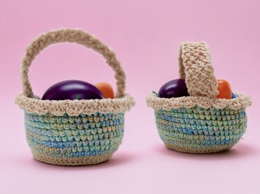 Zwei kleine gehäkelte Körbchen in Türkis- und Beigetönen mit Henkeln, gefüllt mit violetten und orangefarbenen Ostereiern vor rosa Hintergrund.