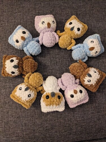 Mehrere kleine gehäkelte Amigurumi-Eulen in Pastell- und Erdtönen, rund auf einem Stoffuntergrund angeordnet