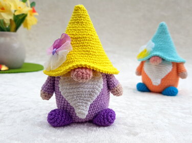 Zwei farbige Amigurumi-Wichtel mit spitzen Hüten und weißen Bärten auf hellem Untergrund, links eine Vase mit Narzissen