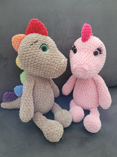 Zwei gehäkelte Amigurumi‑Drachen, beige mit Regenstacheln und rosa mit Wimpern, sitzend