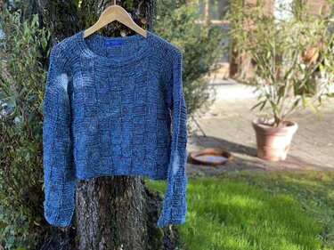 Variegated blue crocheted women's sweater with checkerboard texture hanging on a wooden hanger against a tree trunk