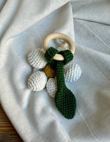 Gehäkelter Baby-Beißring mit Holzring, weißen Häkelelementen, gelbem Zentrum und grünem Blatt auf hellem Stoff