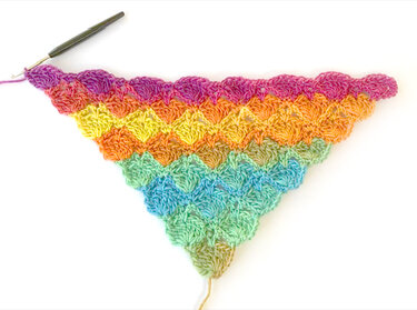 Anfang eines gehäkelten Dreieckstuchs in Regenbogenfarben mit Muschelmuster und Häkelnadel