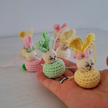 Nahaufnahme: Hand hält kleine gehäkelte Osterhasen in Grün und Gelb mit bemalten Holzköpfen; weitere Hasen unscharf im Hintergrund.