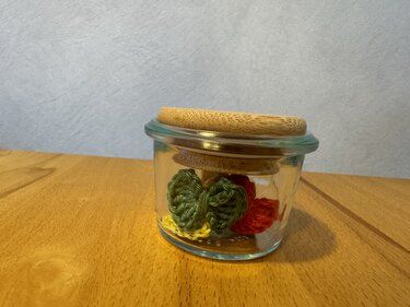 Kleine gehäkelte Herzen in Grün, Rot und Gelb in einem Glas auf einer Holzoberfläche