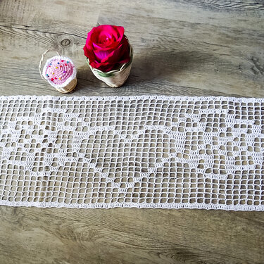 Weißer gehäkelter Tischläufer mit Herzmuster auf hellem Holz, rote Rose und Mini-Cupcake