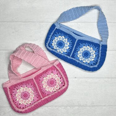 Zwei gehäkelte Umhängetaschen, eine rosa und eine blau, mit weißen Blumenmotiven in je zwei Quadraten und breiten Trägern