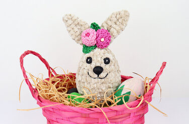 Häkelhase mit rosa Blumenapplikation sitzt in einem pinken Osterkorb mit Bastfüllung und bemalten Eiern