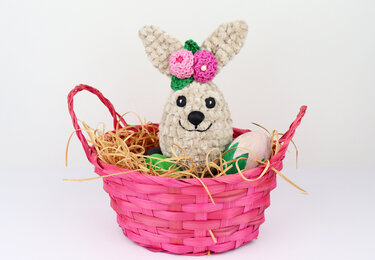 Crocheted bunny with pink flowers sitting in a bright pink woven basket with straw-like filler and decorative eggs.