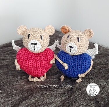 Zwei gehäkelte Amigurumi-Teddybären mit roten und blauen herzförmigen Körpern, beigen Köpfen und weißen Flügeln auf flauschigem Untergrund.
