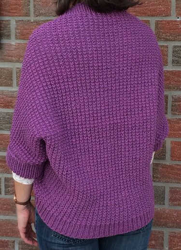 Rückenansicht eines violetten, kurzärmeligen gestrickten Seelenwärmers mit strukturierter Maschenoptik