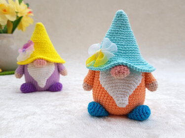 Zwei gehäkelte Amigurumi-Wichtel, vorne orange mit türkisfarbener Mütze und Blüte, im Hintergrund ein lila Wichtel mit gelber Mütze
