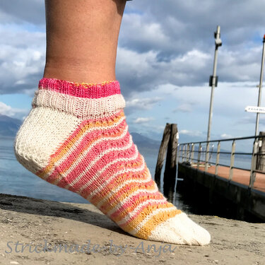 Gestrickte Kurzsocke mit rosa, gelben und weißen Streifen an einem Fuß am Wasserrand