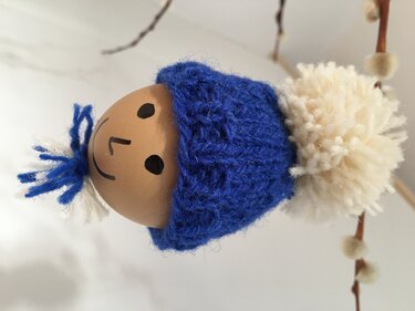 Gestricktes Osterei mit blauer Mütze, aufgemaltem Gesicht und weißem Pompon, aufgehängt an einem Weidenkätzchenzweig.
