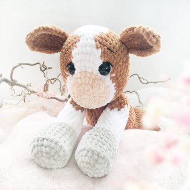 Gehäkeltes braun-weißes Amigurumi-Kalb mit großen Augen liegend auf hellem Untergrund