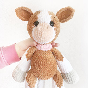 Gehäkelte braun-weiße Amigurumi-Kuh mit rosa Schal, von einer Hand gehalten.