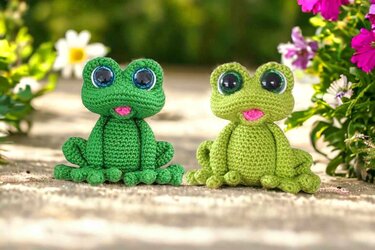 Zwei gehäkelte Amigurumi‑Frösche, links dunkleres Grün, rechts helleres Grün, große Augen, rosa Mund.