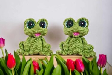 Two green crocheted amigurumi frogs with large glossy eyes and pink mouths, sitting on a wooden board among tulips.