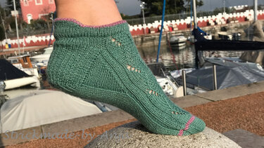 Grüne knöchelhohe Stricksocke mit rosa Bündchen und Lochmuster auf einem Stein vor Booten im Hafen