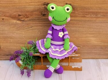 Green crocheted frog in a purple ruffled dress seated on a wooden bench with lavender