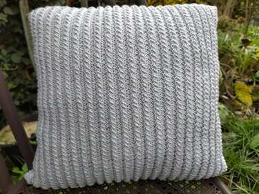 Hellgraues Strickkissen mit vertikaler Rippenstruktur auf einem Stuhl im Garten