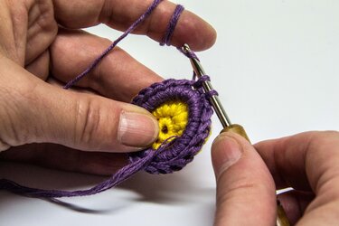 Hände häkeln mit einer Häkelnadel lila Maschen um ein gelbes Häkelstück.