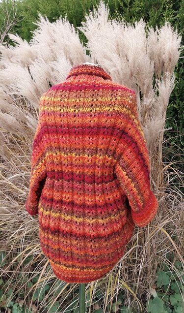 Rückansicht gestrickter Seelenwärmer in Rot‑Orange‑Gelb mit Streifen- und Lochmuster, auf Ständer vor Ziergras