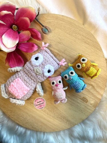 Vier gehäkelte Eulen-Amigurumis in Beige, Rosa, Türkis und Gelb auf einer runden Holzplatte mit rosa Blüte.