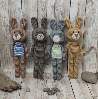 Vier gehäkelte Amigurumi-Hasen in Beige, Braun, Grau und Senf mit kleinen roten Herzen vor rustikaler Holzplanke