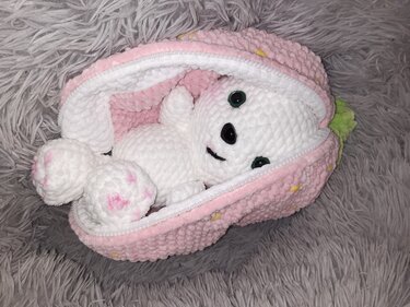 Weißer gehäkelter Amigurumi-Hase mit rosa Akzenten in geöffneter rosa Erdbeertasche auf grauem Flauschuntergrund