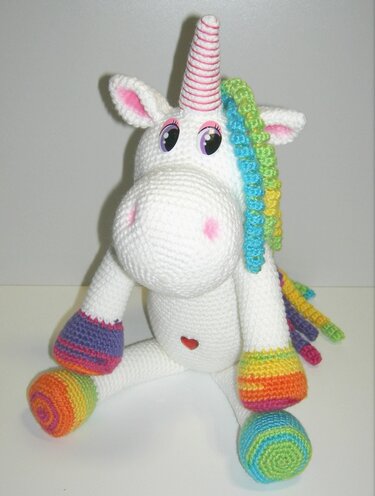 Weißes gehäkeltes Einhorn mit Regenbogenmähne, rosa gestreiftem Horn und bunten Hufen vor neutralem Hintergrund.