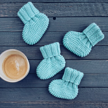 Vier türkisfarbene gestrickte Babyschühchen und eine Kaffeetasse auf dunklem Holz