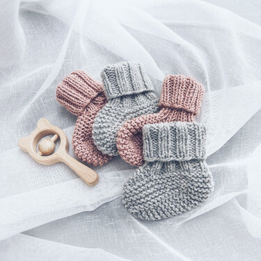 Four hand-knitted baby booties (two grey, two pink) with a wooden animal rattle on white fabric.