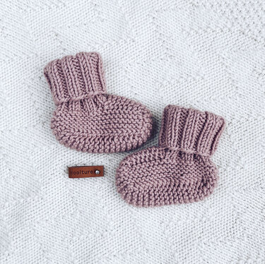 Pair of dusty pink baby socks showing garter stitch soles and folded ribbed cuffs on a white textured blanket