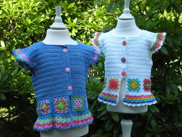 Zwei gehäkelte Kinderwesten (blau und weiß) mit Granny-Squares und Knöpfen an Schaufensterpuppen.