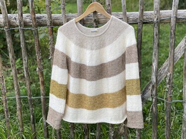 Gestrickter Damenpullover mit breiten Querstreifen in Creme, Beige, Braun und Senfgelb an rustikalem Holzzaun hängend.